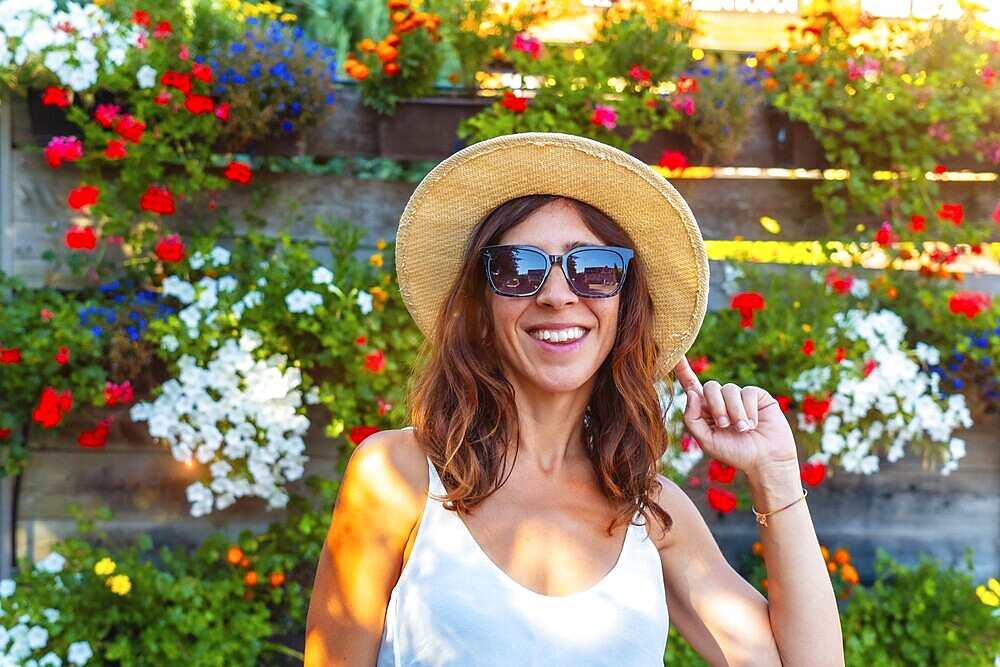 A woman wearing a straw hat and sunglasses is smiling in front of a wall of flowers. The flowers are in various colors and are arranged in a way that creates a beautiful and vibrant display