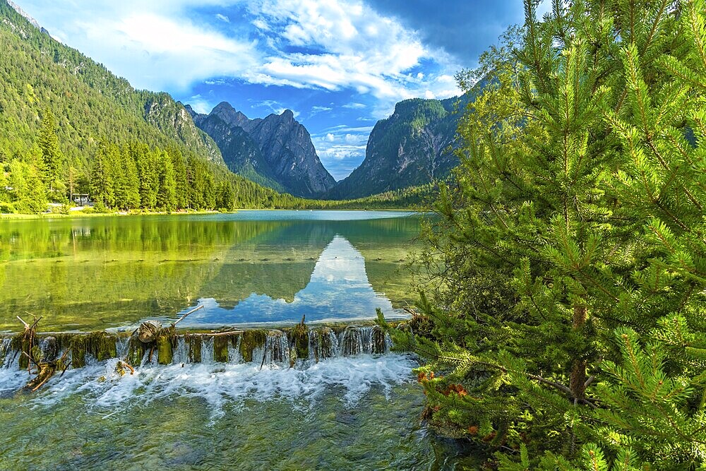Crystal clear water cascading from a small waterfall into lake dobbiaco, reflecting the majestic dolomite peaks under a summer sky