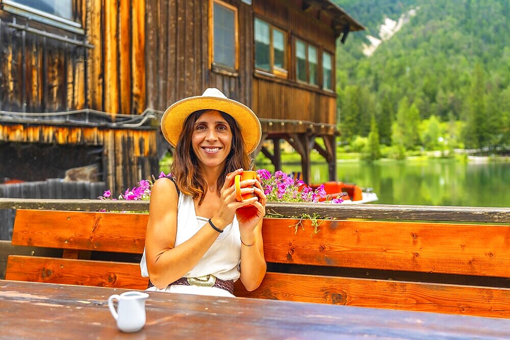 Woman with straw hat enjoying a hot drink in a bar with mountain lake view in the dolomites