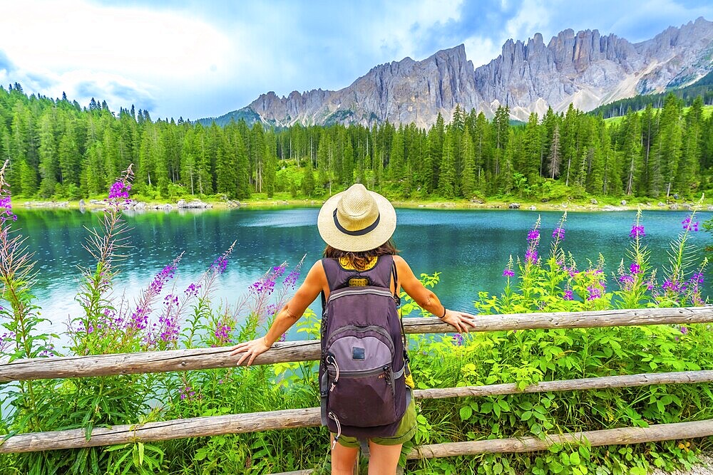 Female tourist with backpack admiring the stunning turquoise waters of lake carezza and the latemar mountain range in the italian dolomites during summer