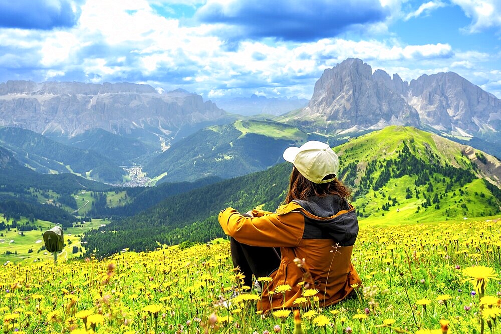 Woman hiker sitting in a vibrant flower field, admiring the stunning seceda peaks rising majestically in the dolomites of italy