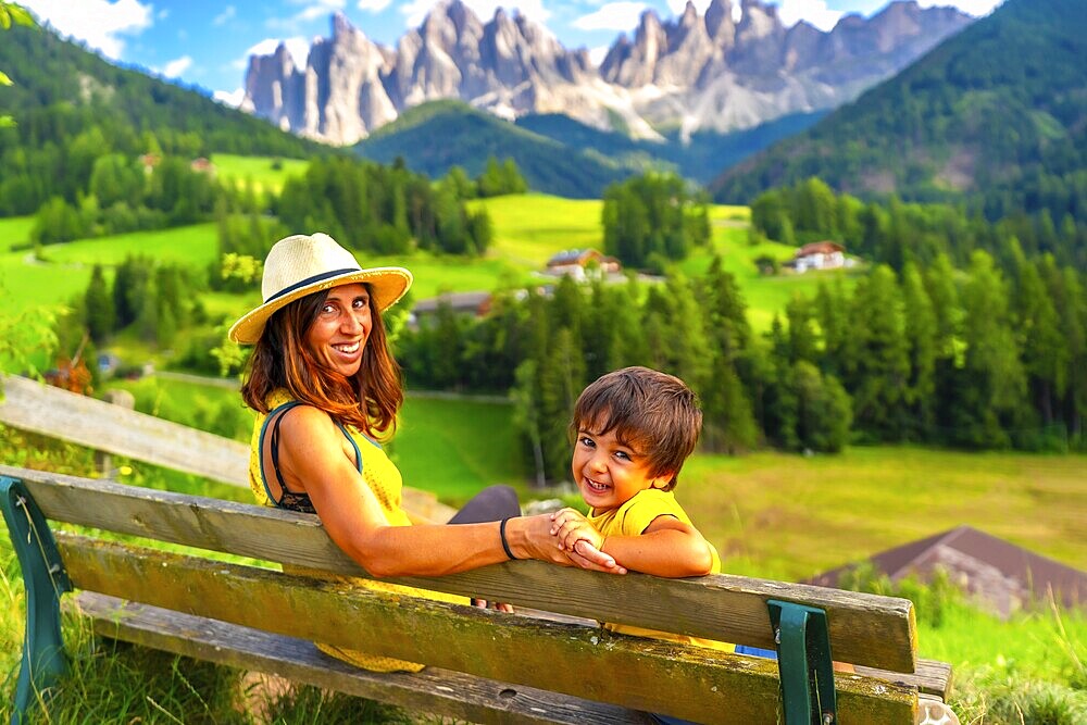 Mother and son sitting on a bench, admiring the breathtaking landscape of the italian dolomites near santa maddalena church in val di funes