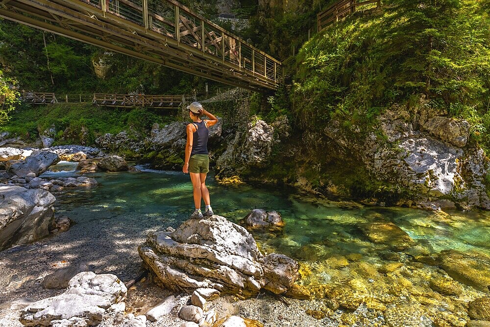 Female tourist standing on rock admiring tolmin gorges with wooden bridge, crystal clear turquoise water and lush vegetation in summer