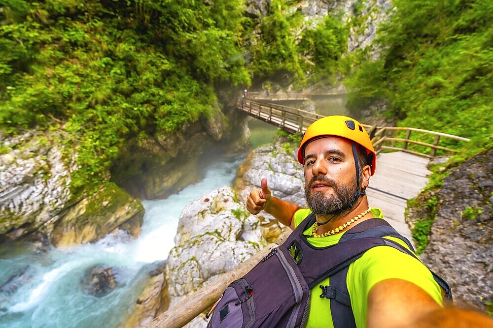 Tourist wearing helmet and backpack hiking in vintgar gorge, near bled, slovenia, making thumbs up sign by the radovna river