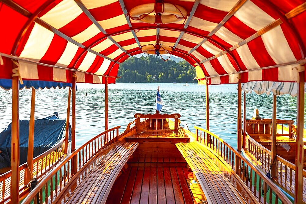 Wooden tourist boat with red and white canopy sailing on lake bled in slovenia, offering scenic view of bled island