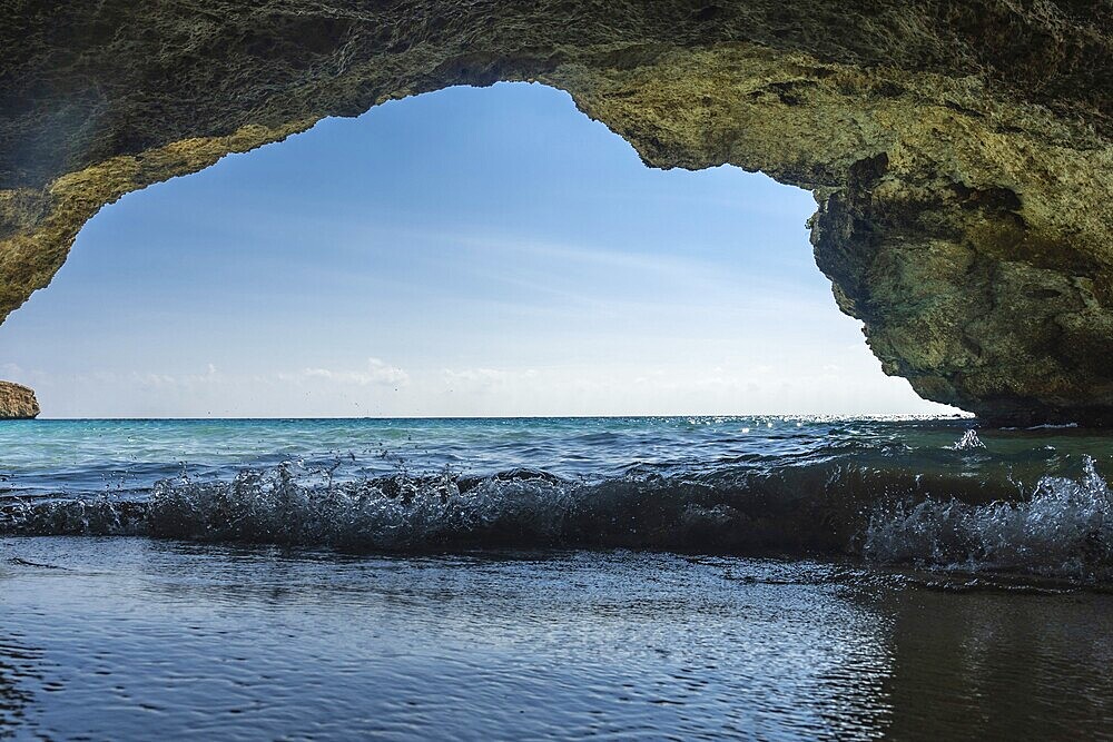 Gentle waves lapping at the shore, viewed from beneath a rocky overhang on a sunny beach in cala domingos, mallorca