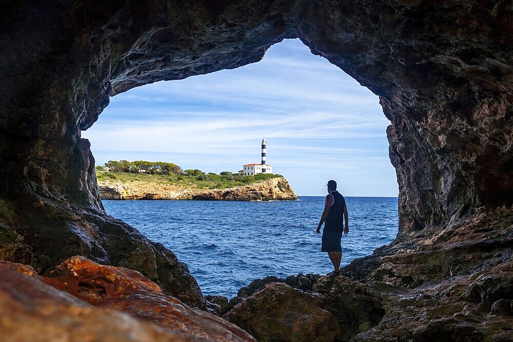 Tourist stands inside the arc natural cave in portocolom, mallorca, admiring the scenic view of a lighthouse across the blue waters