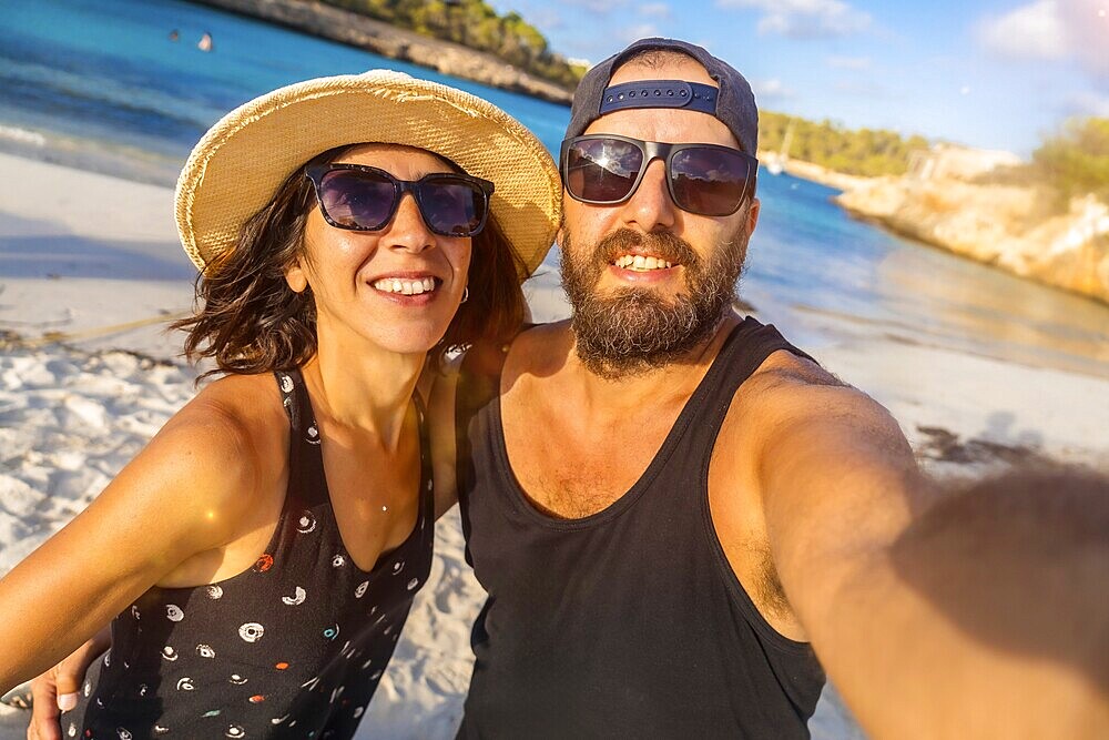A man and woman are smiling for the camera on a beach. The man is wearing a black tank top and sunglasses, while the woman is wearing a straw hat