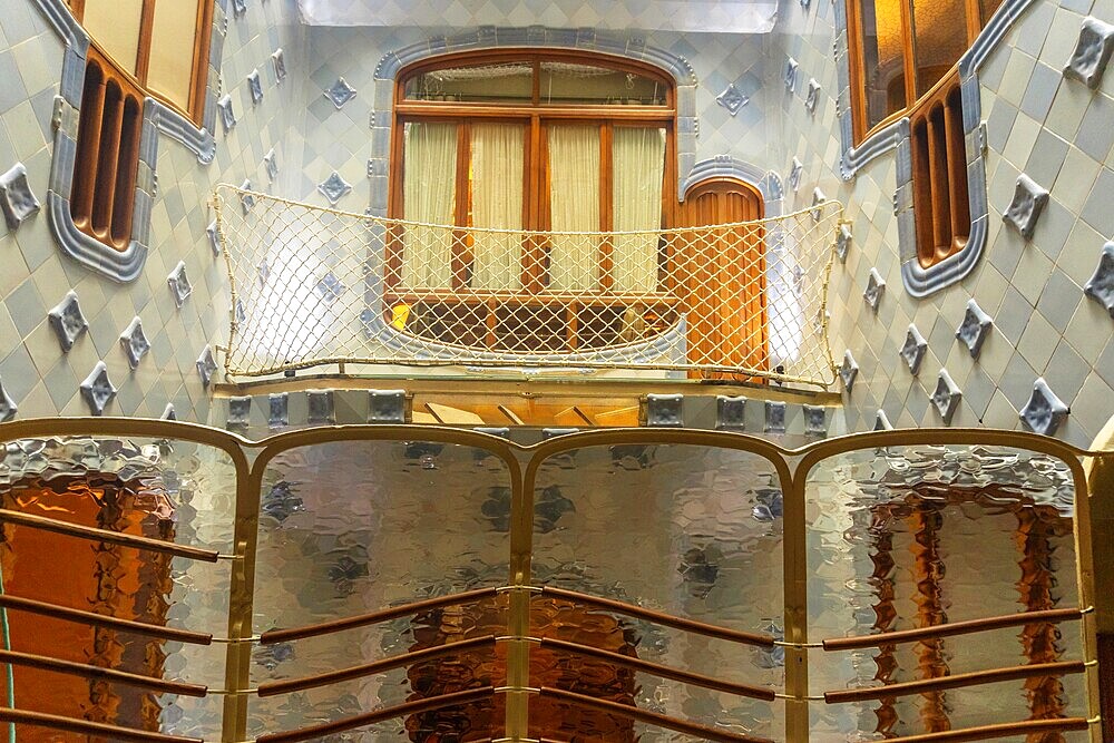 Light filtering through textured glass panels illuminates the unique blue tiled walls and wooden balcony of casa batllo, showcasing antoni gaudi's architectural brilliance