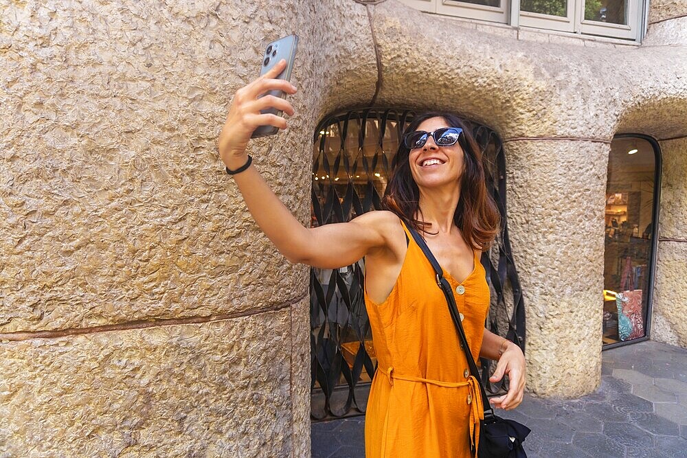Smiling woman taking selfie with smartphone in front of casa mila in barcelona, enjoying summer tourism in catalonia