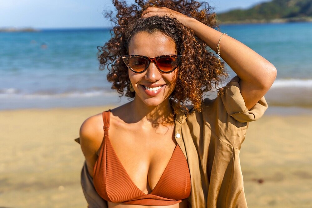 Sensual woman in bikini and hands on hair smiling at camera wearing sunglasses at beach