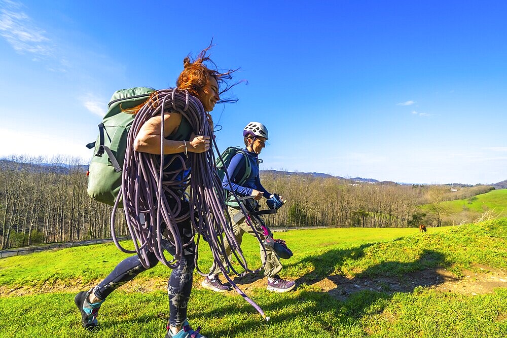 Two female climbers are walking across a mountain meadow carrying climbing rope and gear on a sunny day