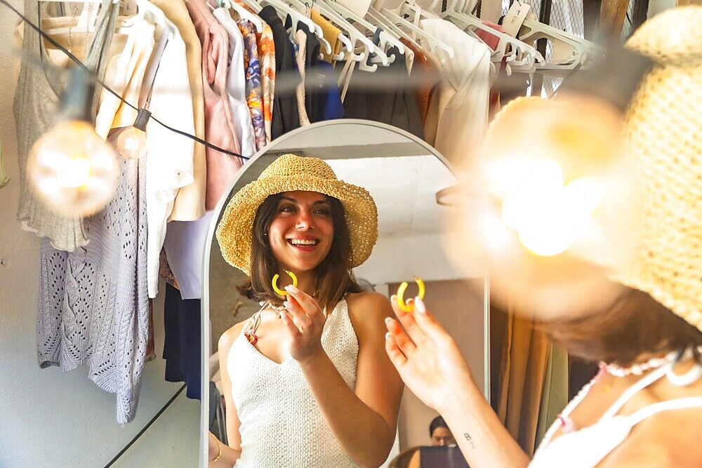 Young woman wearing straw hat trying on yellow earrings, getting ready in front of a mirror in her hotel room or bungalow in siargao island, philippines
