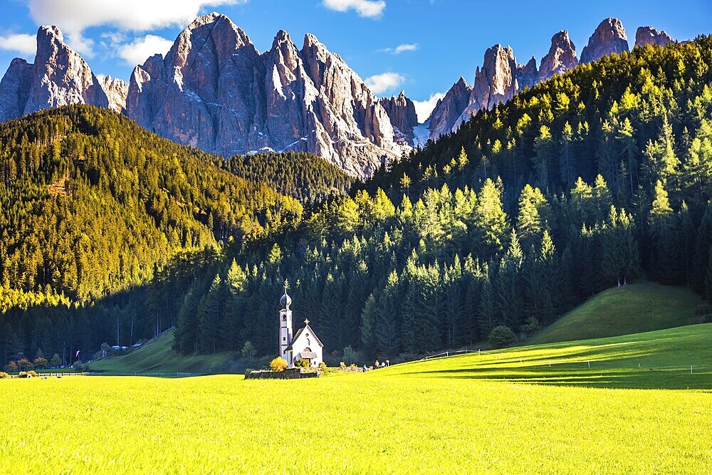 Dolomites, Tirol. Sunny autumn day. The famous symbol of the valley Val di Funes - church of Santa Maddalena. Rocky peaks and forested mountains surrounded by green Alpine meadows