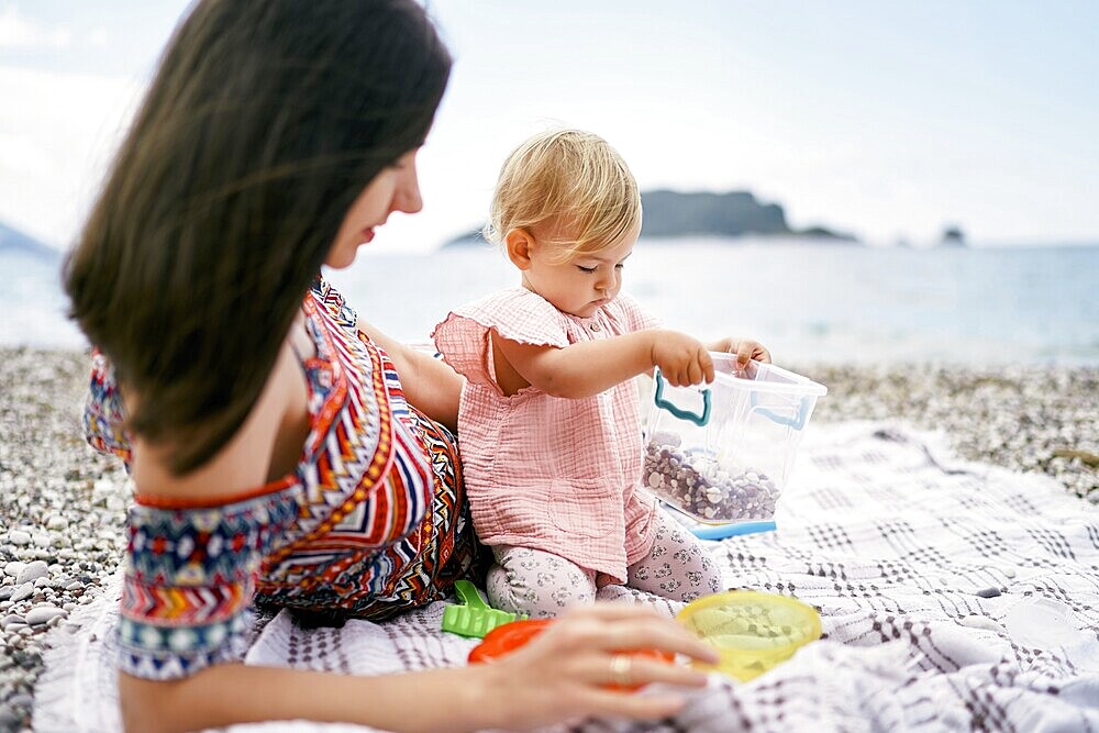 Mom lies on a blanket on a pebble beach next to a sitting little girl.