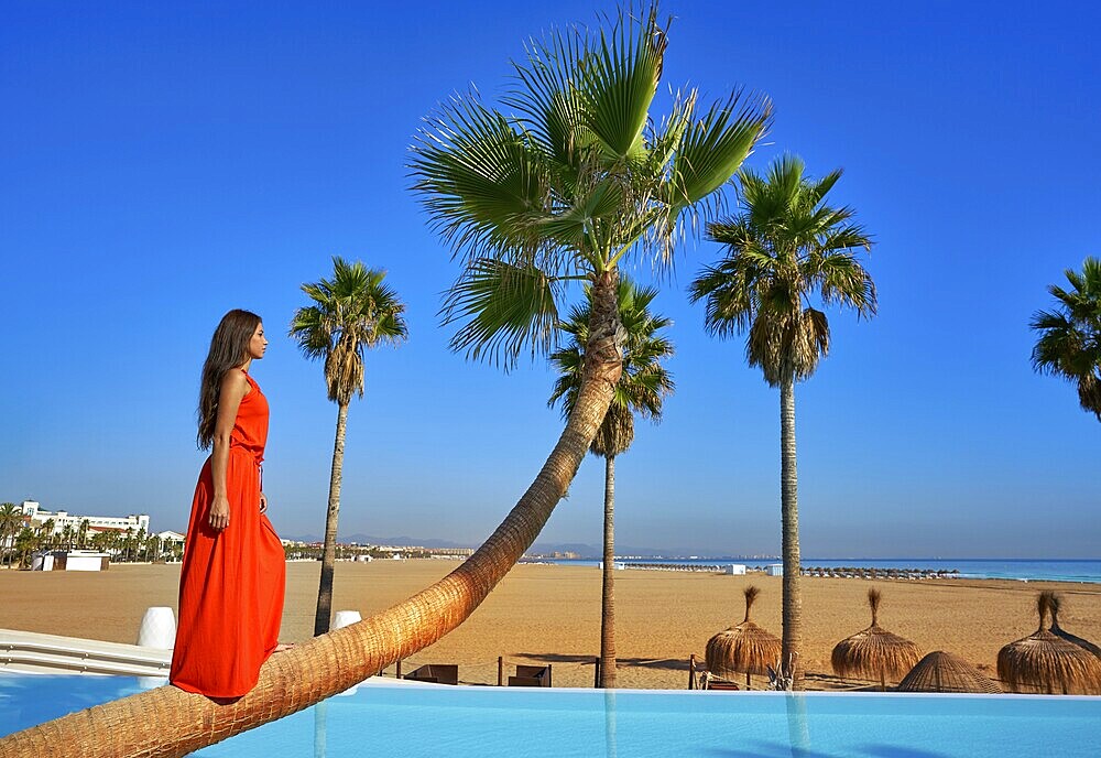 Beautiful woman standing on bent palm tree trunk at infinity pool long red dress