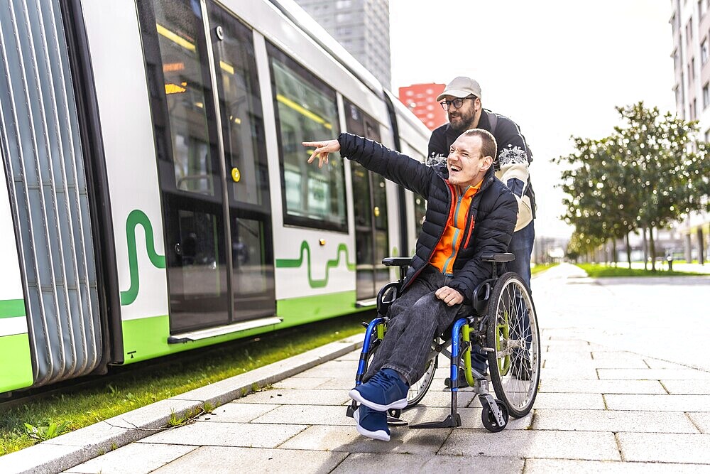 Disabled man in a wheelchair points at something while his friend stands behind him near a tram
