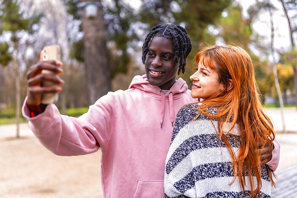 Young multiracial couple smiling together while capturing a joyful selfie with a smartphone in a vibrant park setting