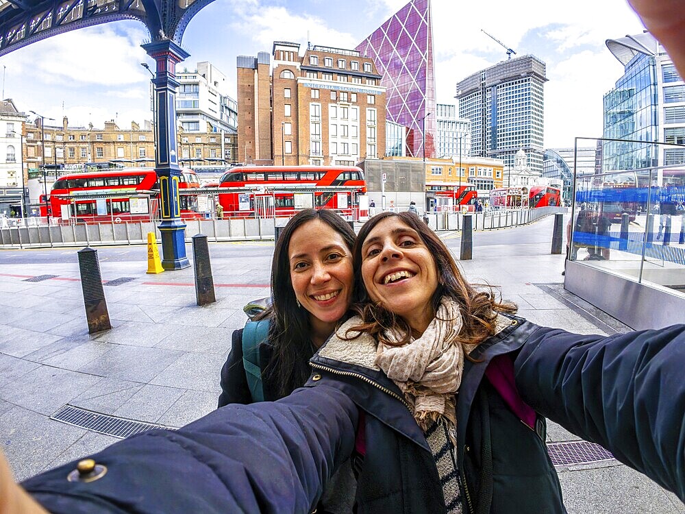 Two happy tourists taking a selfie in the city of london, with red buses and modern buildings in the background