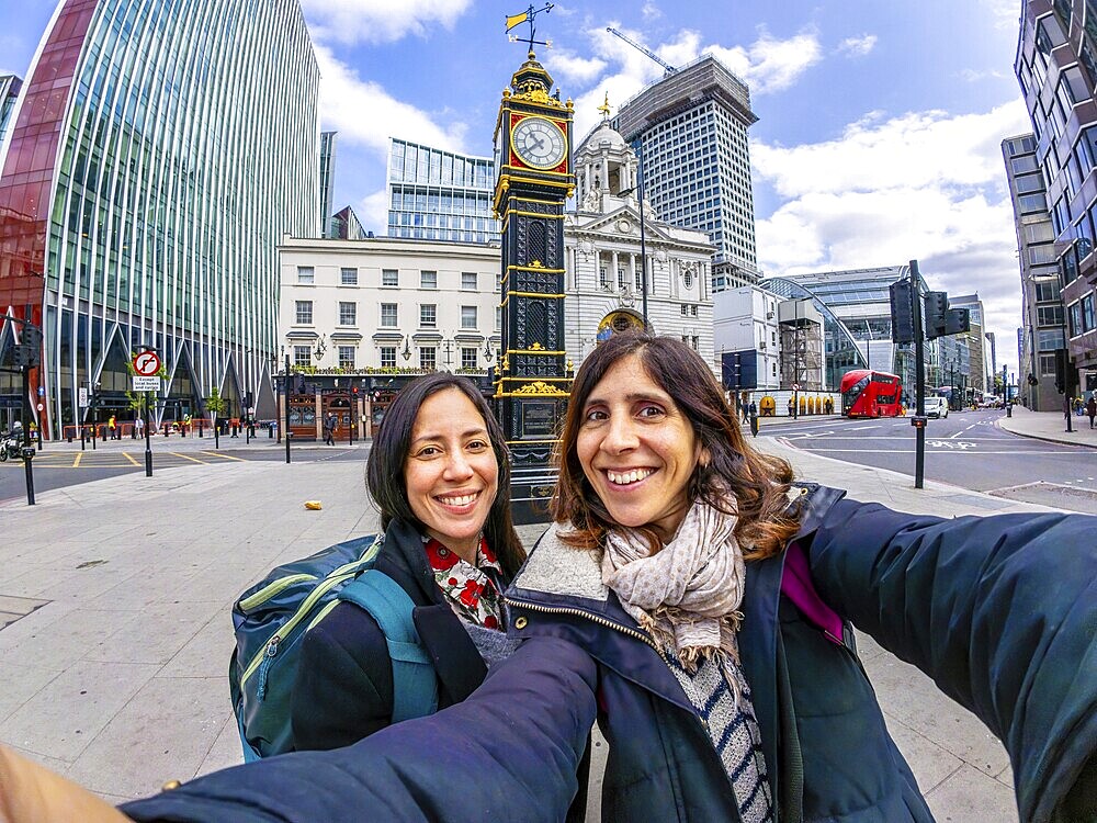 Two happy tourists taking a selfie in london near a historical clock tower, with modern buildings and red bus in the background