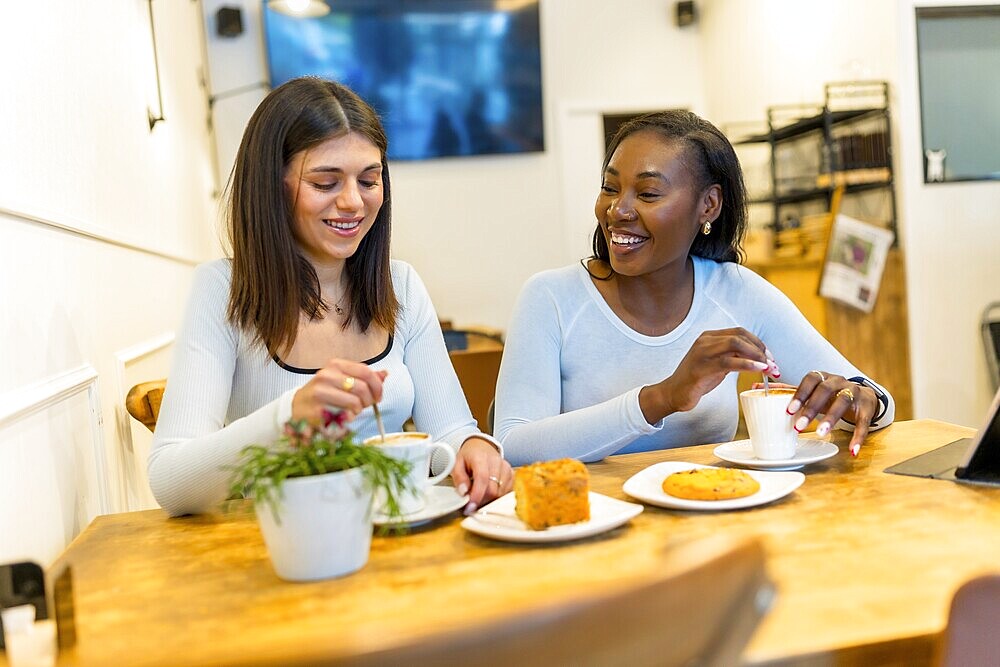 Two young women enjoying a coffee break, chatting and laughing together in a vibrant, modern cafe filled with natural light