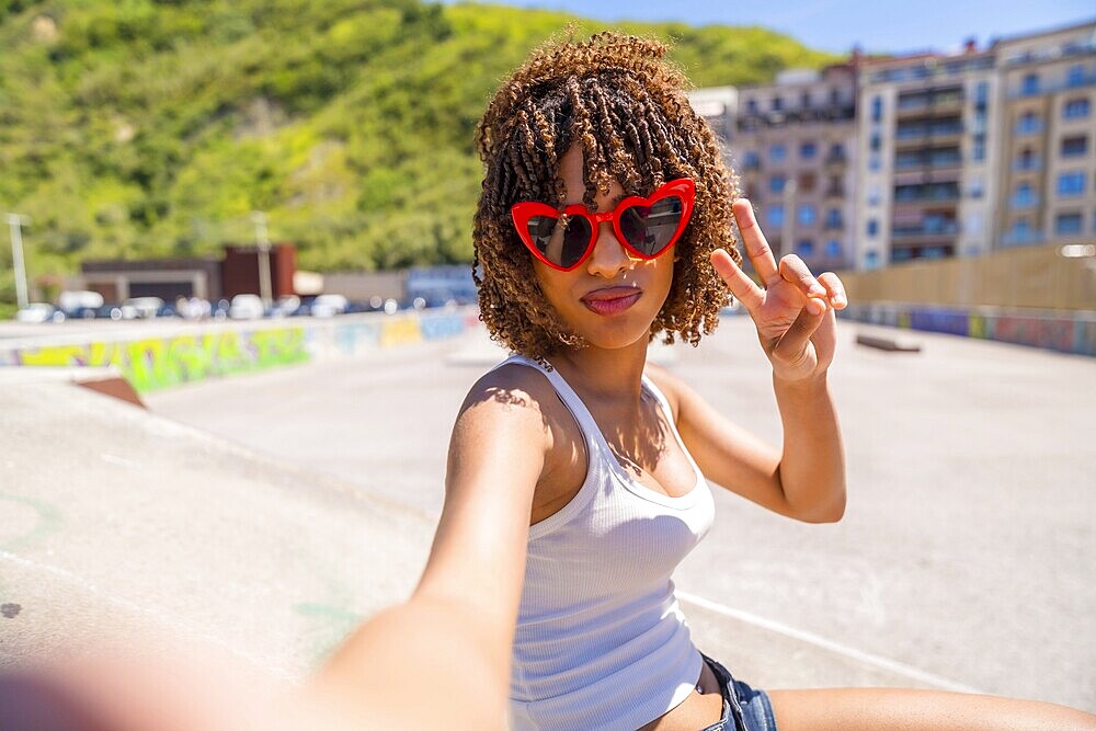 Fashionable young woman with heart shaped sunglasses making peace sign while taking a selfie in skatepark
