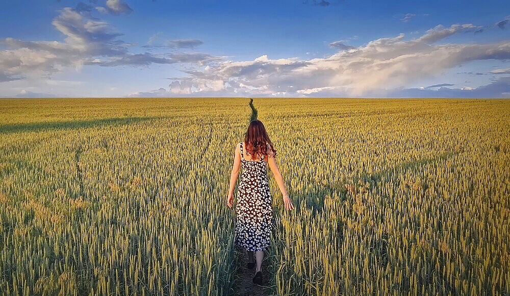 Carefree woman walking a pathway across a green wheat field. Idyllic countryside summer scenery, freedom concept