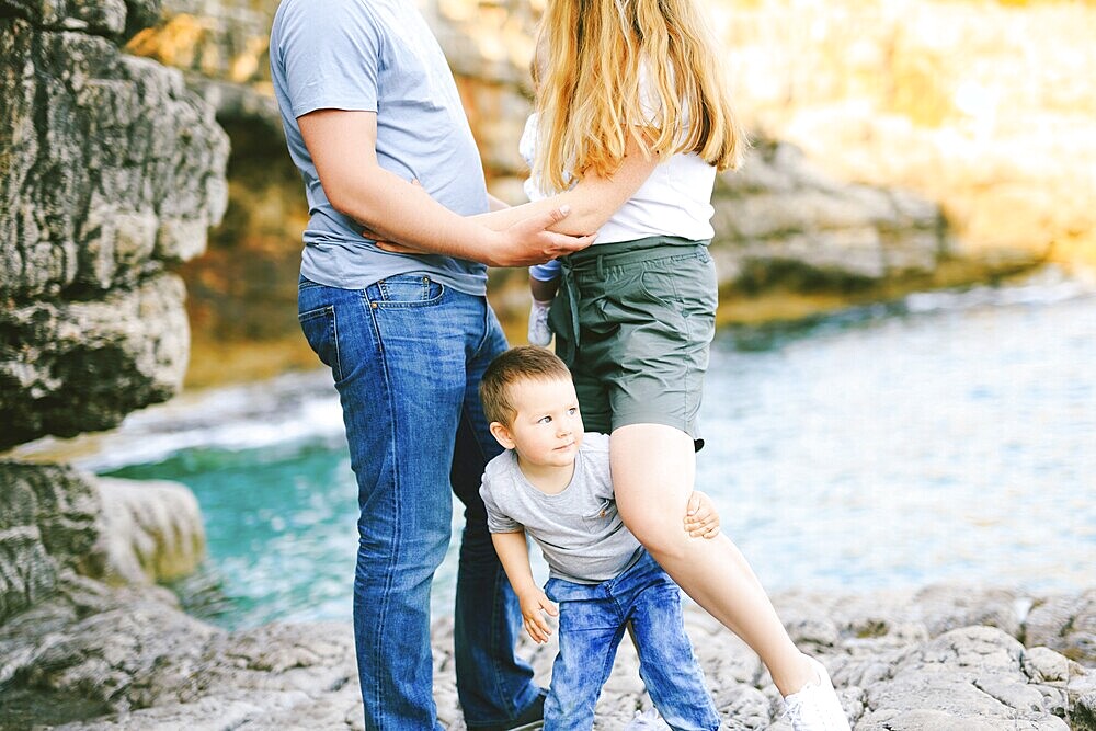 Mom and Dad stand on the beach, holding hands, against the backdrop of the sea and rocks. Little boy holds his mother's leg and looks out from behind her.
