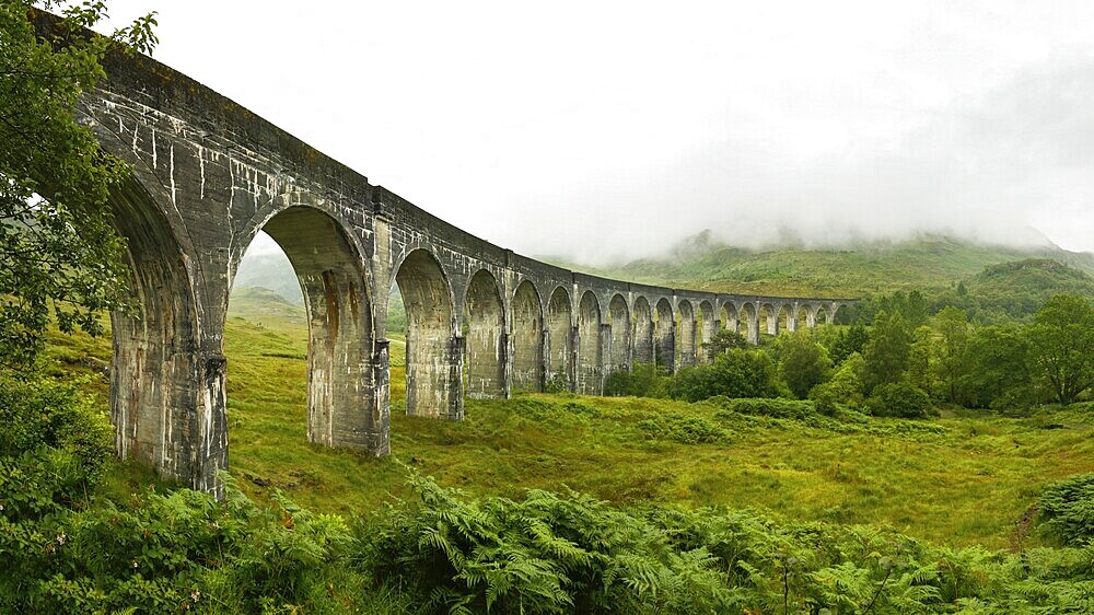 High resolution panorama of Glenfinnan railway viaduct (location from Harry Potter movie) shot from side, on overcast day with grey sky and lot of green grass and trees around. Inverness, Scotland, United Kingdom