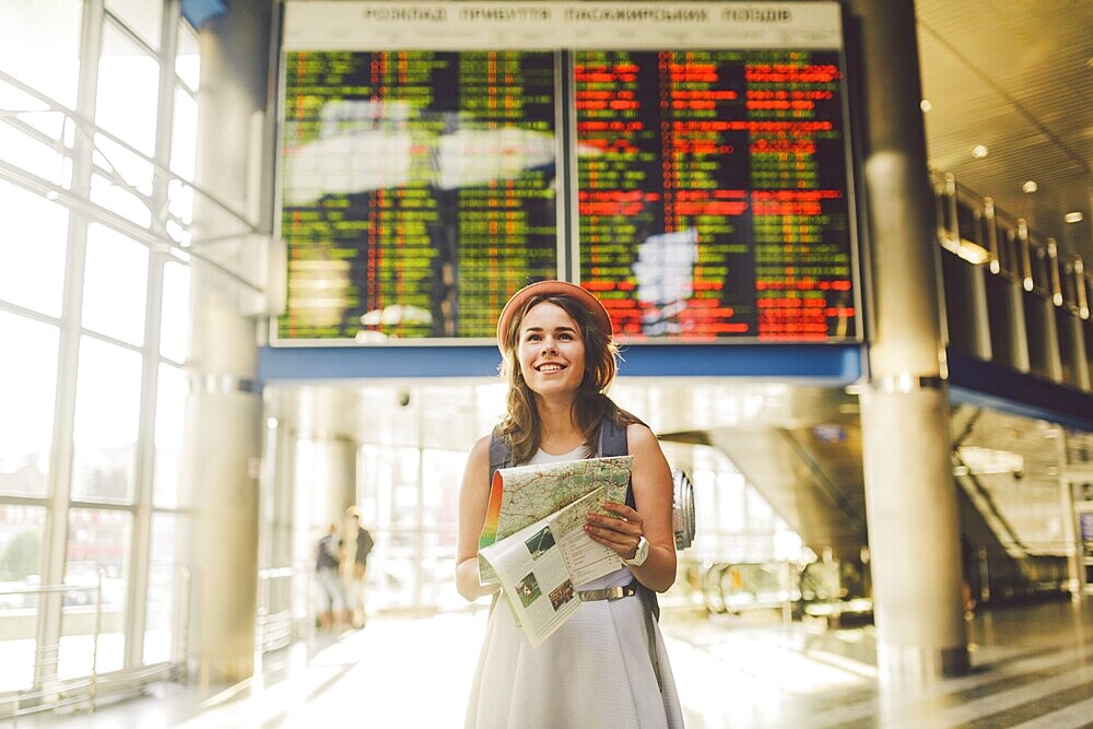 Theme travel and transportation. Beautiful young caucasian woman in dress and backpack standing inside train station terminal looking at electronic scoreboard holding phone, map paper hand navigation