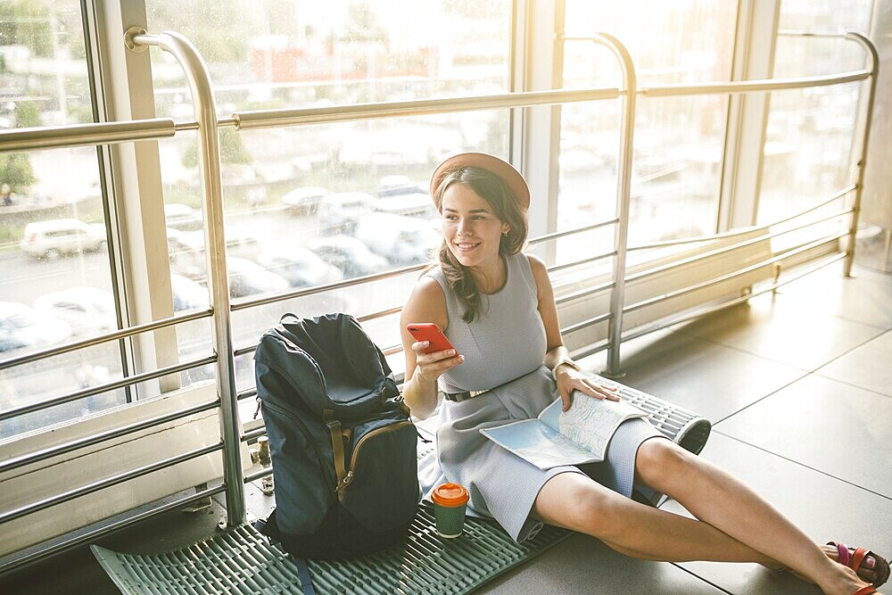 Theme of tourism and travel of young student. Beautiful young caucasian girl in dress and hat sits on floor tourist rug inside terminal airport terminal. Waiting room delayed flight, delay departure
