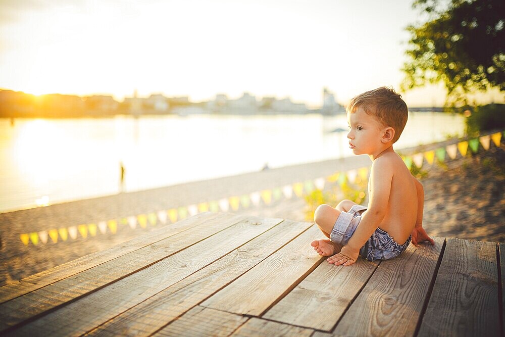 The theme is child and summer beach holiday. A small Caucasian boy sits sideways on a wooden pier and admires the view of the sandy beach and the pond, the river. With bare legs in blue denim shorts