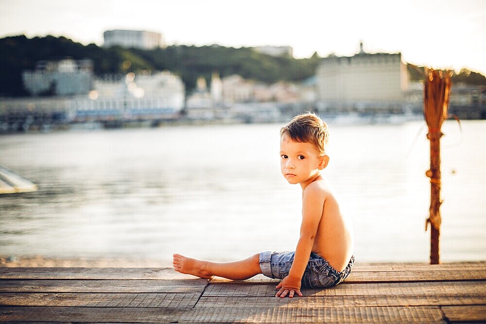 The theme is a child and summer beach holiday. A small Caucasian boy sits sideways on a wooden pier and looks at the camera on a sandy beach and a pond, a river. With bare legs in blue denim shorts