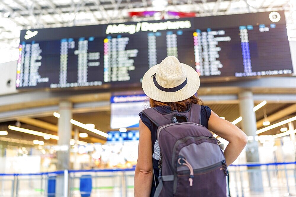 A woman wearing a straw hat and a backpack is standing in front of a large airport display. She is waiting for her flight, possibly at an airport