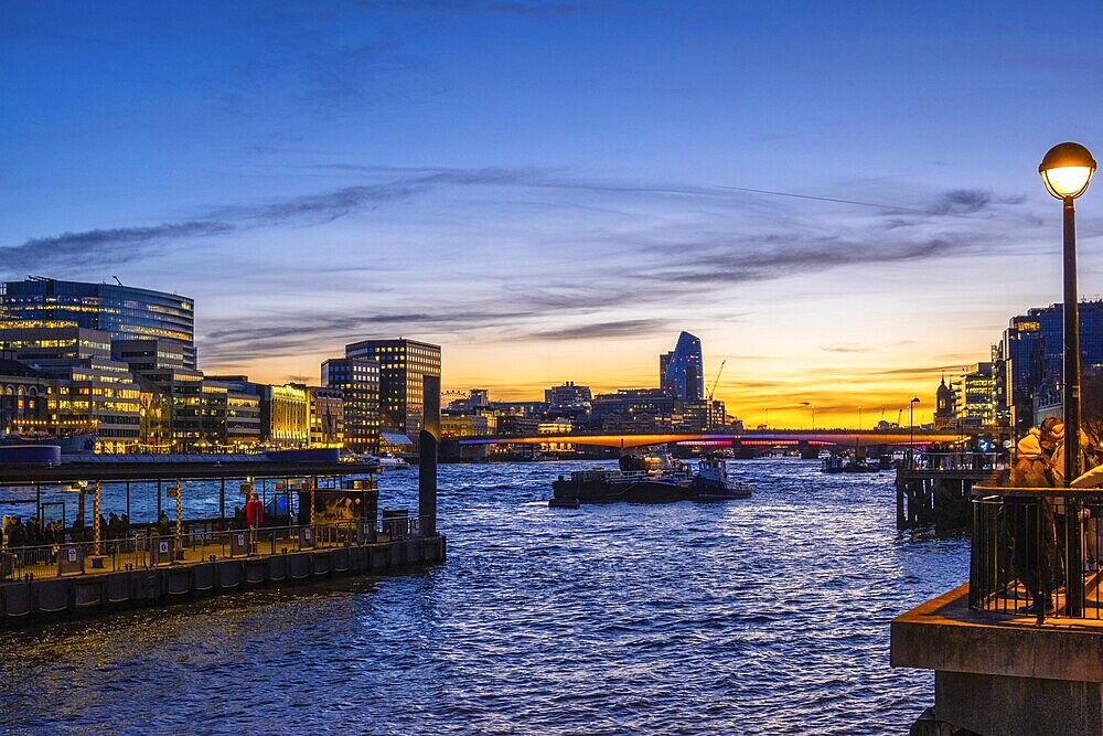 Tugboats sailing on thames during sunset with London’s illuminated skyline reflecting on the water
