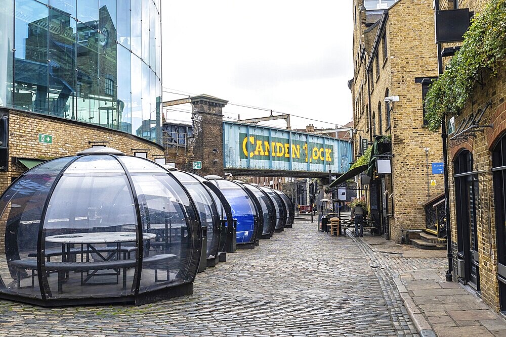 Transparent dining pods line a cobblestone street beneath the Camden lock bridge in London, offering a unique and sheltered dining experience
