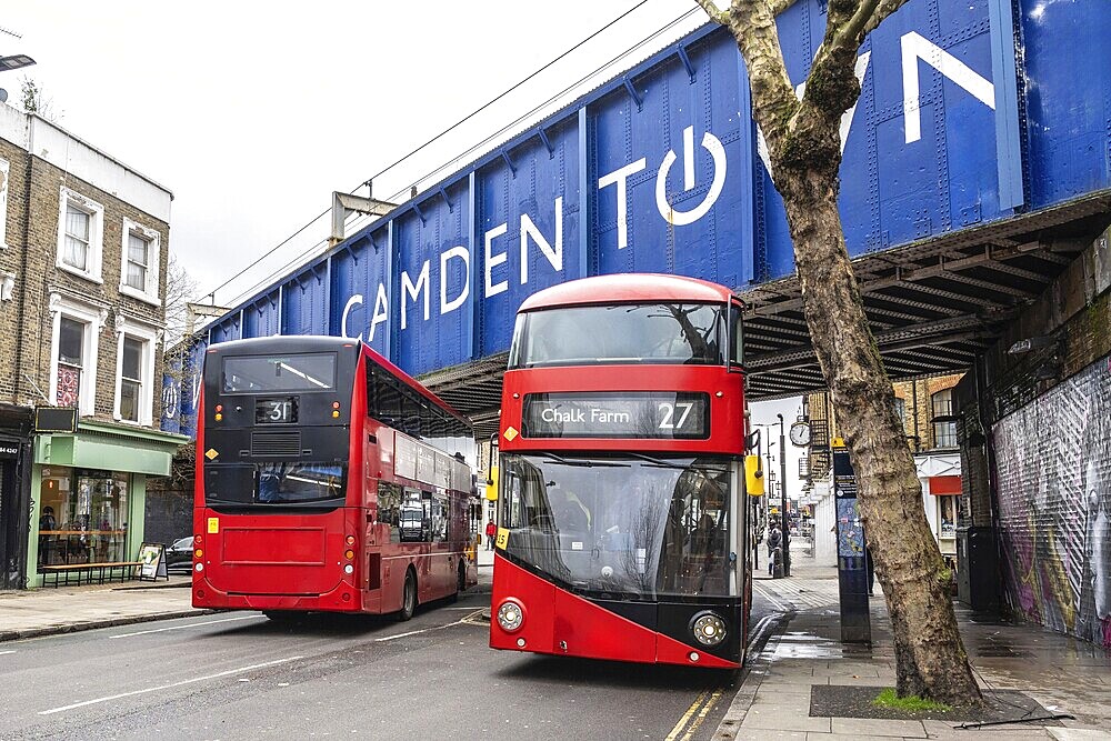 Red double decker buses driving on a London street, passing under a bridge with the Camden town sign
