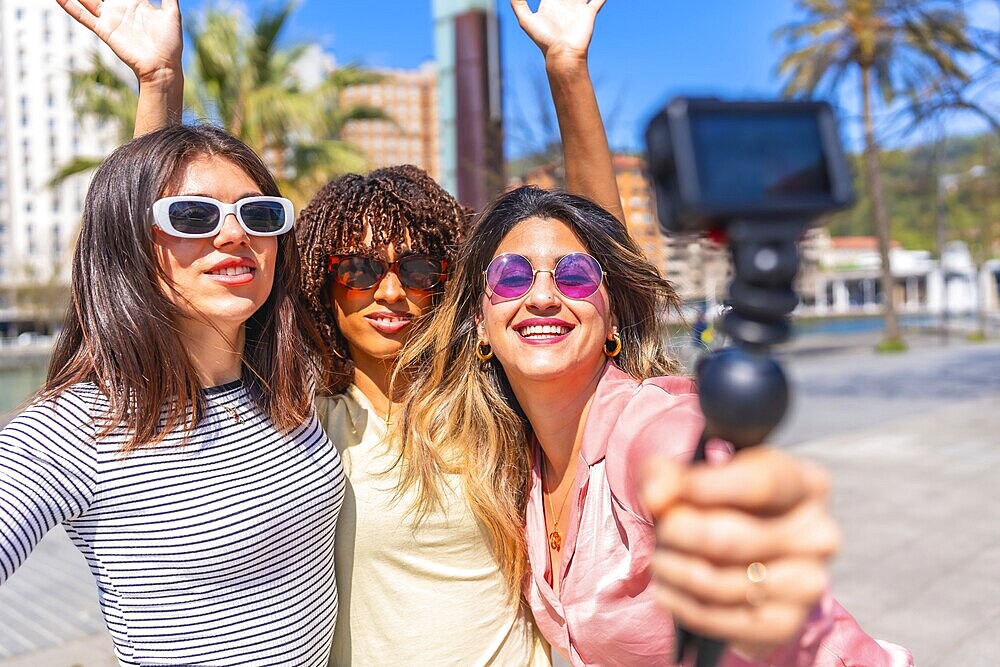 Three cheerful women taking a selfie with action camera, waving and smiling, enjoying a summer day together in the city
