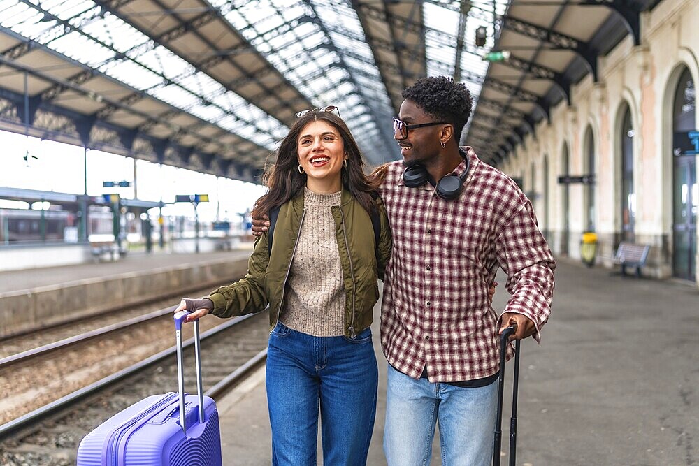 Young multi ethnic couple walking together with luggage at a train station platform, enjoying their vacation