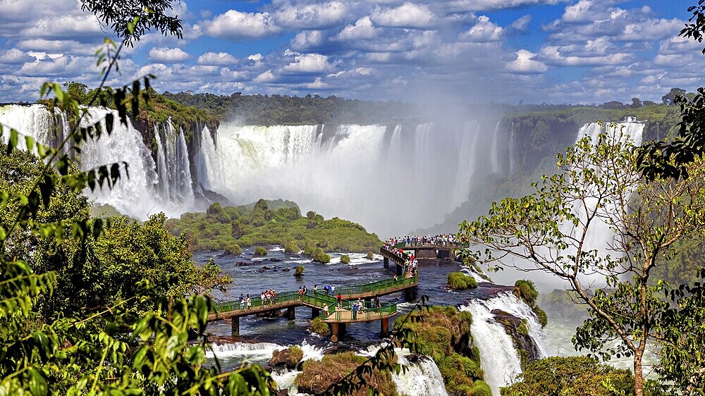 View of a lively waterfall and tourists on a bridge, surrounded by lush greenery and clouds, The Iguazu Falls between Argentina and Brazil