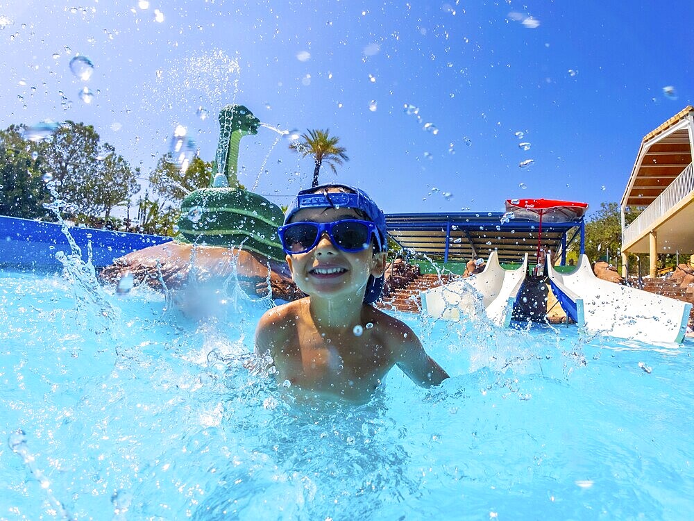 Young boy wearing blue sunglasses, joyfully splashing in a swimming pool at a vibrant water park, embracing summer fun and adventure