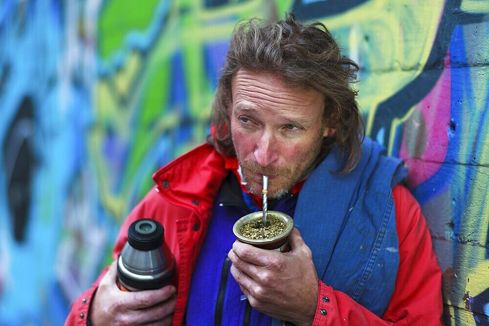 A man with a beard in colourful urban surroundings drinks mate from a cup. Relaxed atmosphere in front of a graffiti wall, portrait shoot, Unterallgäu district, Bavaria, Germany