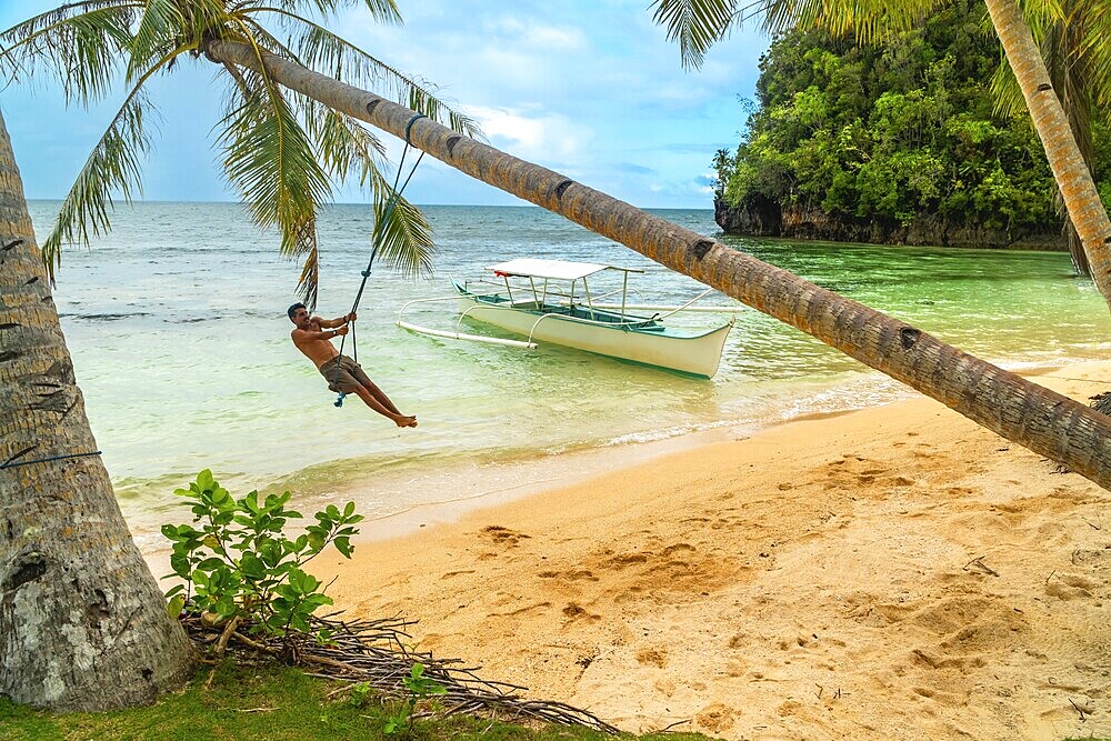Tourist enjoying a rope swing beneath palm trees on a tropical beach with traditional outrigger boat on kangbangyo island, siargao, philippines