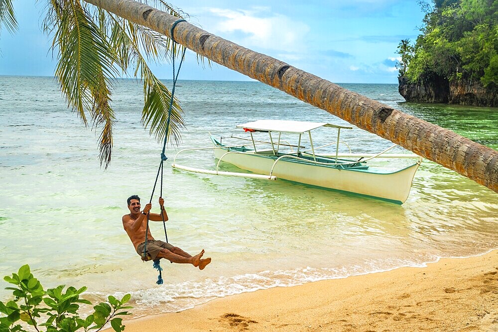 Tourist enjoying a swing on a palm tree with a banca boat on the background, on a beautiful beach in kangbangyo island, siargao, philippines