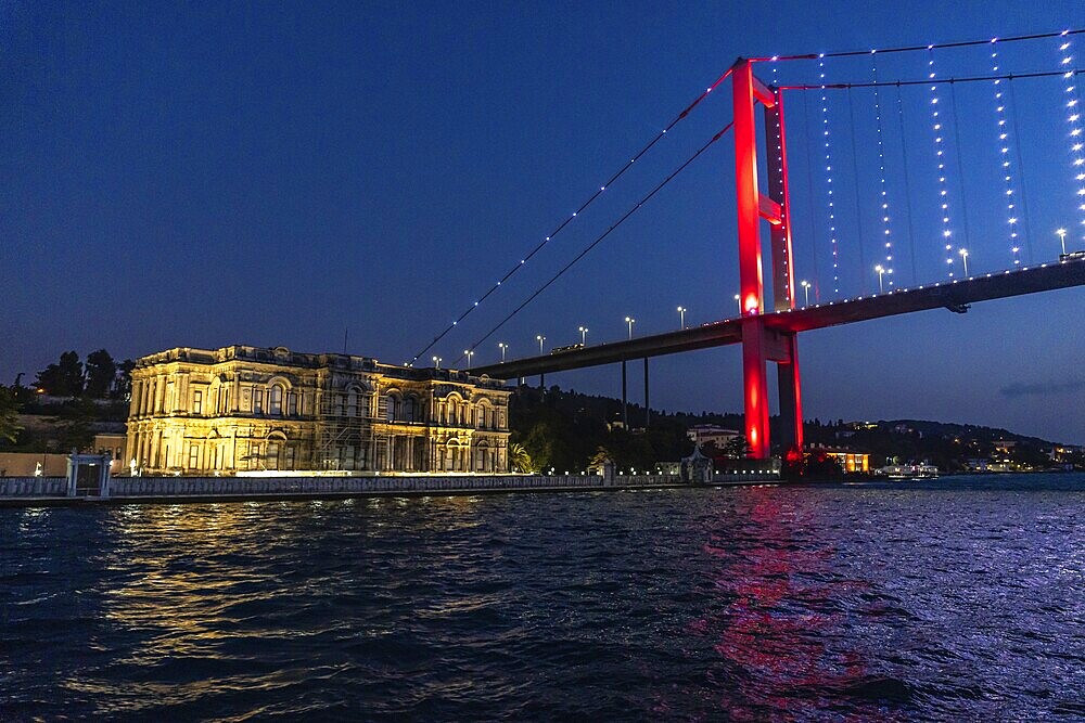 The illuminated suspension bridge over bosphorus strait paints the istanbul night sky red, creating a vibrant contrast with the historic palace
