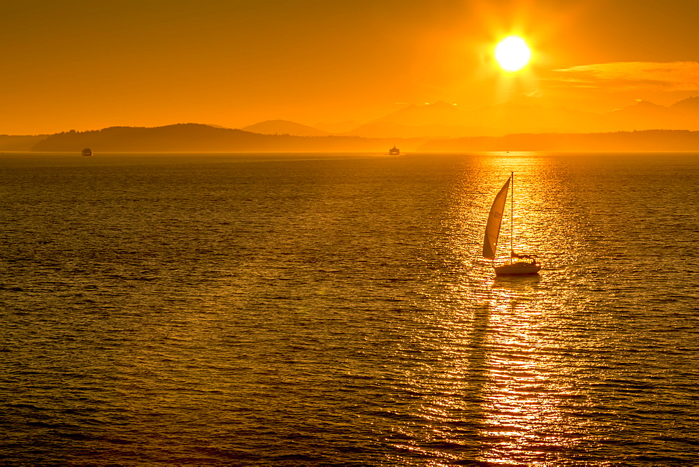 Sailing boat and sunset over Elliott Bay with Bainbridge Island visible on the horizon viewed from Bell Harbour Marina. Seattle, Washington State, United States of America, North America