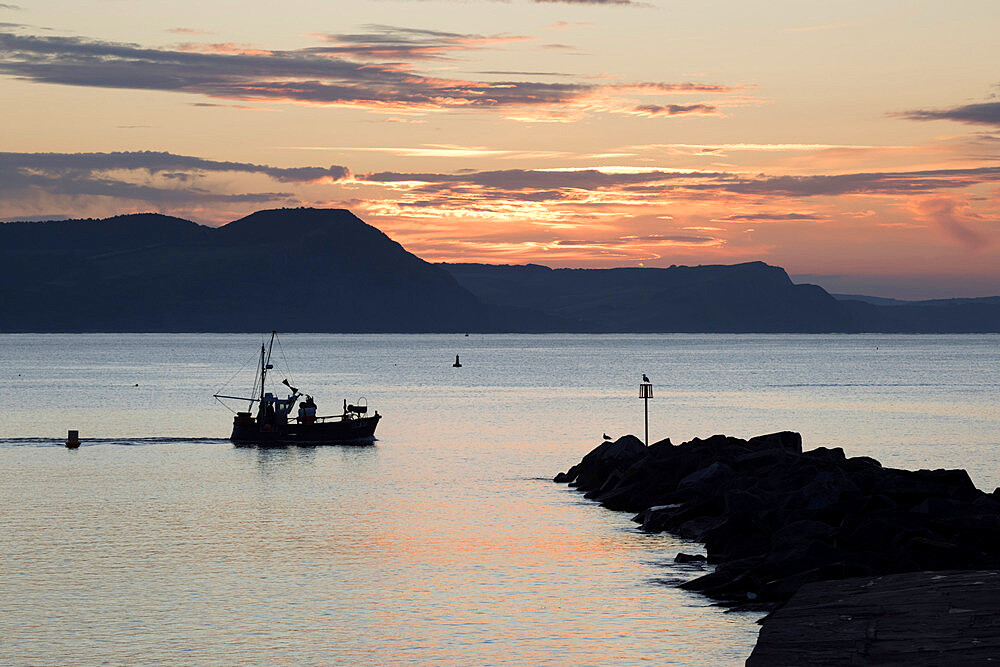 Fishing boat sailing past The Cobb at sunrise with Golden Cap and Jurassic Coast, UNESCO World Heritage Site, Lyme Regis, Dorset, England, United Kingdom, Europe