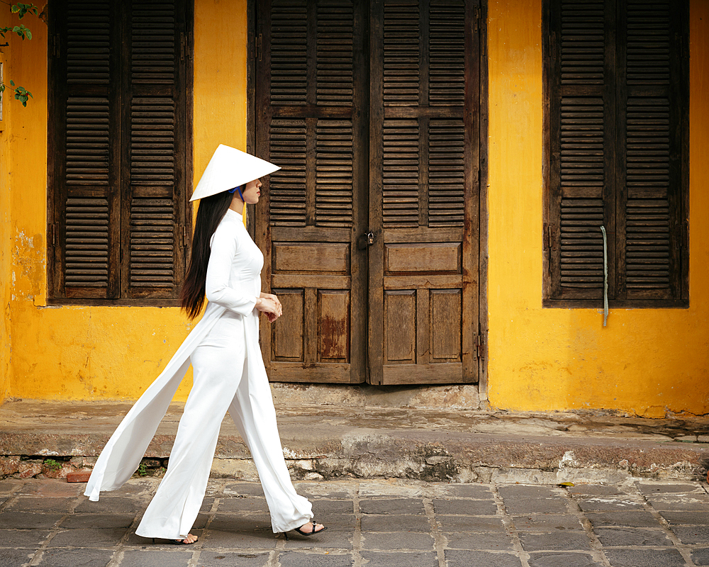 Vietnamese woman in ao dai walking past buildings, Hoi An, Quảng Nam Province, Vietnam