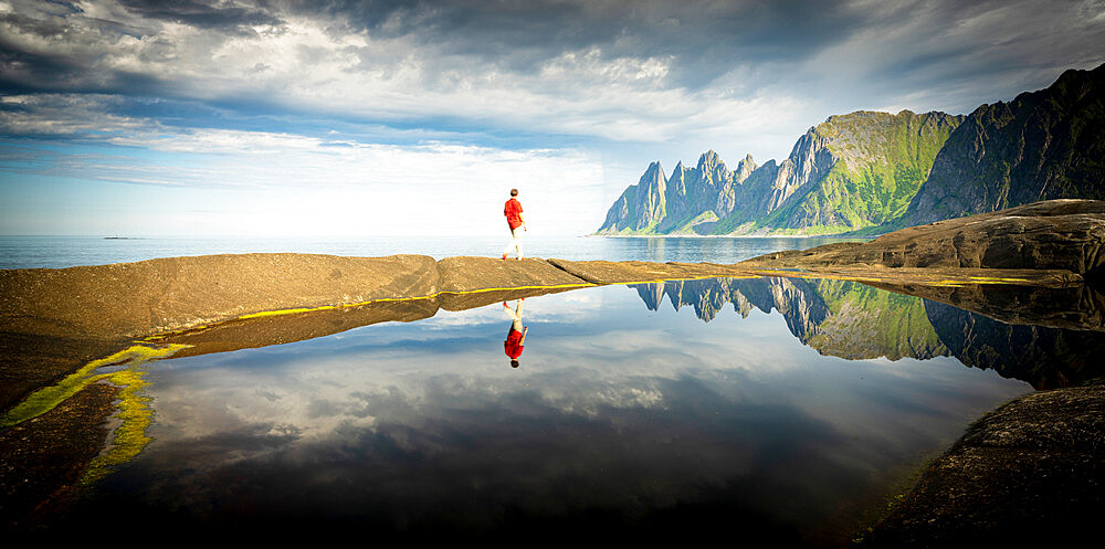 Hiker man walking on water's edge admiring mountains reflected in the sea, Tungeneset, Senja, Troms county, Norway, Scandinavia, Europe