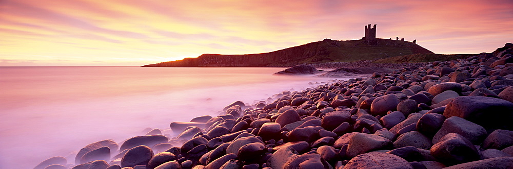 An imposing silhouette of Dunstanburgh Castle against a magnificent sky at sunrise with a beach of basalt boulders in the foreground, Embleton Bay, near Alnwick, Northumberland, England, United Kingdom, Europe